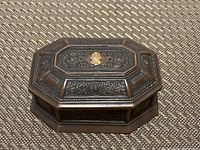 Closed coffin-shaped jewelry box showing embossed floral patterns, gold accent emblem in the center, and octagonal shape