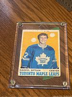 Photo of Darryl Sittler rookie hockey card in protective case on wooden surface, showing card wear and player portrait in Leafs uniform.