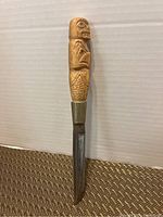 Full view of the cutting utensil showing the carved wooden handle with Indigenous-style figures and the metal blade.