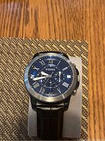 Photo showing a men's Fossil wristwatch with a round blue dial, Roman numerals, three subdials, silver-tone case and black leather strap placed on a textured surface.