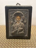 Front face of silver religious icon showing detailed raised depiction of a haloed saint figure inside an ornate frame on black plaque.