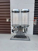 Front view of double cereal dispenser with clear cylindrical containers and chrome components on a gray surface against a concrete background.