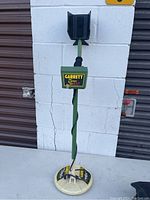 Green Garrett Super Scanner GS-100 metal detector standing upright against a white cinder block wall with a brown garage door seen in background.