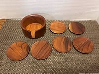 Photo showing six round teak wood coasters arranged on a mat with the teak holder bowl behind them. Coasters have smooth finish and visible wood grain.