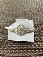 Paratrooper badge on a textured background, showing front design with parachute and wings in silver metal.