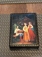 Top lid of the box showing a detailed hand-painted scene of three people in traditional Russian clothing in front of a cabin, surrounded by gold decorative border.