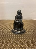 Front view of a small silver-colored statue showing a man seated with one leg crossed and arm resting on knee, detailed face and long hair visible.