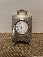 Front view of sterling silver rectangular travel clock case with inset clock face on white dial, Arabic numerals, and small seconds dial.