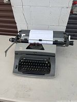 Gray manual Smith Corona SCM typewriter with black keys on a white surface. Paper loaded in typewriter. Visible brand and model markings.