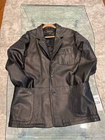 Full front view of a men's black leather dress coat laid flat on a rug, showing the button closure and pockets.