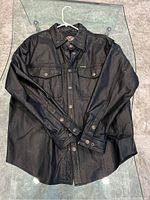 Full view of men's black leather shirt jacket from Harley Davidson, size L, laid flat on glass surface, showing collar, long sleeves, front flap pockets and button closure.