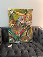Two museum wrap canvas art pieces propped on a black tufted sofa, showing bicycle handlebars with orange tape and a drink glass on a decorative table on one canvas, the other with teal bicycle frame and red drink bottle and glasses.