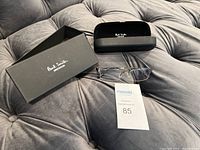 Paul Smith spectacles centered on fabric surface with black open hard case behind and outer matte black box beside it, also includes Maxsold auction card.