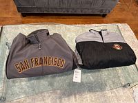 Two folded men's jackets on glass coffee table, front jacket has large 'SAN FRANCISCO' lettering, second jacket folded shows black and gray reversible side with embroidered patch.