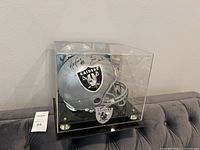 Full-size silver Raiders helmet on black base inside acrylic case