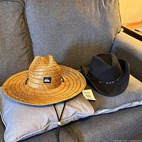 Two men's hats placed on a grey cushion on sofa: black western cowboy hat and tan Quiksilver woven hat with chinstrap.