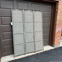 Front view of the three-panel room divider against a brown garage door, showing the neutral woven pattern and hinges.