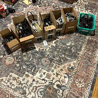 Seven boxes and a green basket arranged on a patterned carpet, filled with assorted phone holsters, chargers, data cables, power cables, adapters, and Christmas lights in used condition.