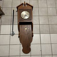 Full view of the antique pendulum wall clock showing the wooden case, pendulum, and clock face.