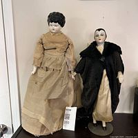 Photo showing both dolls side by side standing upright, Victorian doll on the left with beige dress and Seymour Mann doll on the right with black coat.