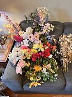 Overview of multiple faux flower bunches laid out on a grey couch, showing a variety of colors including pink, white, yellow, red, purple, and orange flowers along with clusters of dried beige grasses.