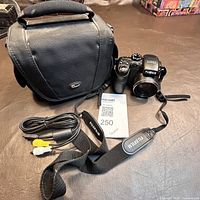 Photo of the Fujifilm Finepix 1000 camera with strap, black fabric carrying case, and AV cable arranged on a dark surface.