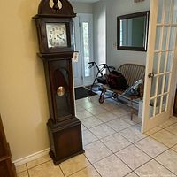 Full view of the antique Mauthe grandfather clock standing in a room showing wooden case, clock face with Arabic numerals and pendulum.