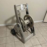 Front angled view of the beige plastic hose reel caddy with wheels and crank handle, showing handle and hose reel spool.