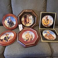 Five John Wayne collector plates in frames displayed on grey fabric sofa. Various frames in round, octagonal and square shapes with wood finish.