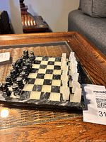 Full view of the miniature chess set showing black and white carved pieces arranged on a checkered board on a glass table.