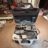 Open gray plastic carrying case containing the vintage Sanyo video camera and accessories including battery, charger, cables, and lens.