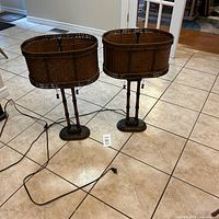 Two rattan style table lamps with oval woven shades and bamboo-like wooden stands shown on tiled floor with cords visible.