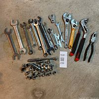 Collection of various hand tools including wrenches, ratchets, sockets, pliers on concrete floor, showing overall lot condition and content.