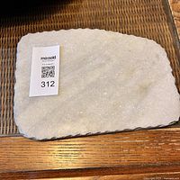 Top view of rectangular stone serving board with rough cut edges placed on a wooden surface with a lot identifier tag on top.