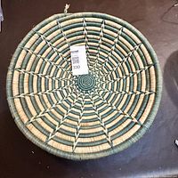 Full view of circular tightly woven coil basket showing star pattern, natural beige and teal colors, hung for photo on dark background.