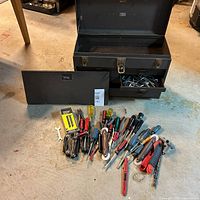 Full view of large black metal vintage tool chest with hinged top lid open showing empty storage, and three small drawers with miscellaneous Allen keys and other small tools inside. Several hand tools spread out in front of chest on floor including screwdrivers, pliers, wrench and rotary cutter with wooden handle.