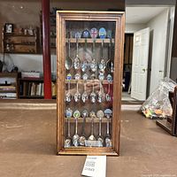Front view of wooden display case with glass door closed showing 18 souvenir spoons