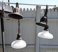 Two antique hanging lamps suspended outdoors showing metal fixtures with patina, white glass shades with scalloped edges, and attached pull chains.