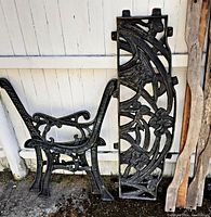Side view of one cast iron bench frame and a wooden guide piece leaning against a wall outdoors.