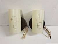 Two cylindrical plastic wall lamps with cut-out atomic star designs and brown backing plates shown from front view with power cords coiled.