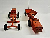 Front view of two vintage orange metal toy vehicles: road grader with blade and dump truck with large rear hopper