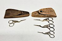 Two sets of antique sewing scissors shaped like storks, one set with one pair and the other with three pairs, all laid flat with their respective cases showing wear and fading.