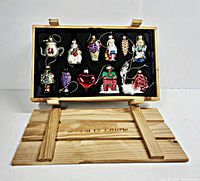 Photo of nine colorful glass Christmas ornaments with various shapes including teapot, snowman, grapes, Santa, pinecone, heart, house, rose, and small figurines arranged in a wooden crate marked David & Laurie.