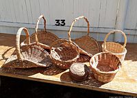 Seven woven baskets displayed on a bench outdoors against a white wooden background, showing all farmer baskets and the sewing basket.