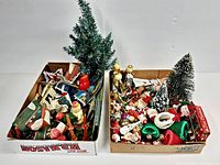 Two cardboard boxes filled with assorted vintage Christmas decor. Left box contains small artificial trees, cardboard houses, and various ornaments. Right box contains more ornaments including elves, Santas, and angels.