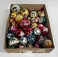 Top-down photo of open cardboard box showing colorful vintage blown glass Christmas ornaments with various painted holiday designs and metallic finishes.