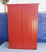 Front closed view of red painted wood cabinet with vertical panel design and crown molding on top.