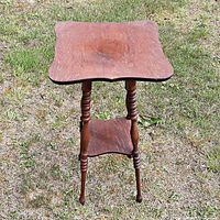 Front angle view of small oak wood side table showing shaped square top, spindled legs, and lower shelf. Visible wear and surface marks on table top.