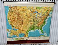 Front view of a large vintage roll-up wall map of the United States with visible wooden rails, showing physical and political features, including Alaska and Hawaii in insets.