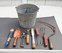 All 8 vintage garden tools and galvanized bucket arranged front-facing on black surface, showing condition and detail of handles and metal parts.